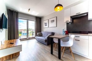 Salon apartamentu z aneksem kuchennym, stołem z krzesłami i wyjściem na balkon.