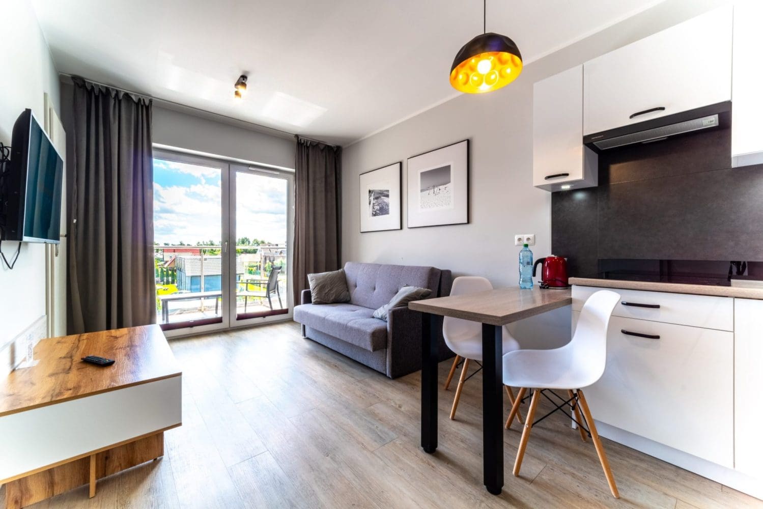 Salon apartamentu z aneksem kuchennym, stołem z krzesłami i wyjściem na balkon.