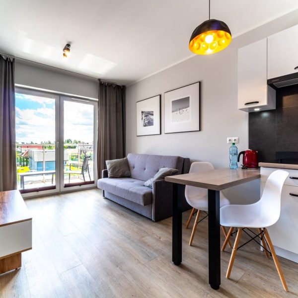 Salon apartamentu z sofą, stołem i aneksem kuchennym, wyjście na balkon
