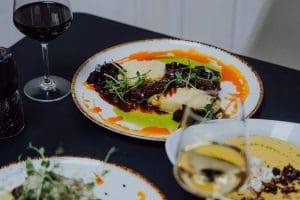 Dwa talerze z rybą na ciemnym stole restauracyjnym, obok kieliszki czerwonego i białego wina.