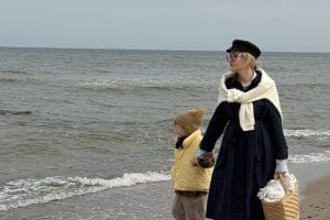 Kobieta w ciemnym płaszczu i dziecko w żółtym kombinezonie na brzegu w pochmurny dzień.