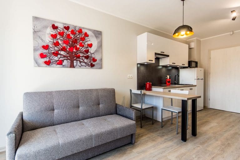 Jasny salon z szarą sofą, aneksem kuchennym i wysokim stołem z krzesłami.