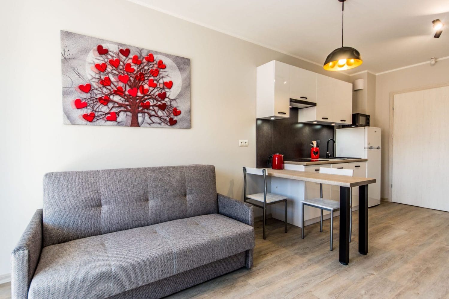 Jasny salon z szarą sofą, aneksem kuchennym i wysokim stołem z krzesłami.