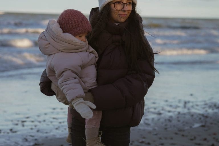 Uśmiechnięta kobieta trzyma na rękach małe dziecko w kurtce i wełnianej czapce, a za nimi fale Bałtyku lśnią w niskim słońcu.