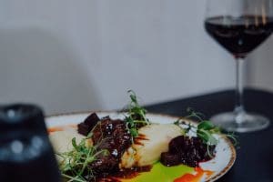 Mięso w ciemnym sosie z puree, buraczkami i kiełkami na ciemnym stole, w tle stoi kieliszek wina.
