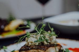 Filet ryby z chrupiącą skórką i kiełkami groszku, a w tle danie mięsne, zupa krem i kieliszek wina.