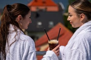 Dwie kobiety w białych szlafrokach z mrożoną kawą na tarasie, w tle czerwone dachy i morze.