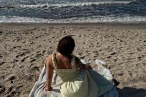 Kobieta w żółtej sukience, widziana od tyłu, siedzi na kocu na szerokiej, słonecznej plaży i patrzy na fale.