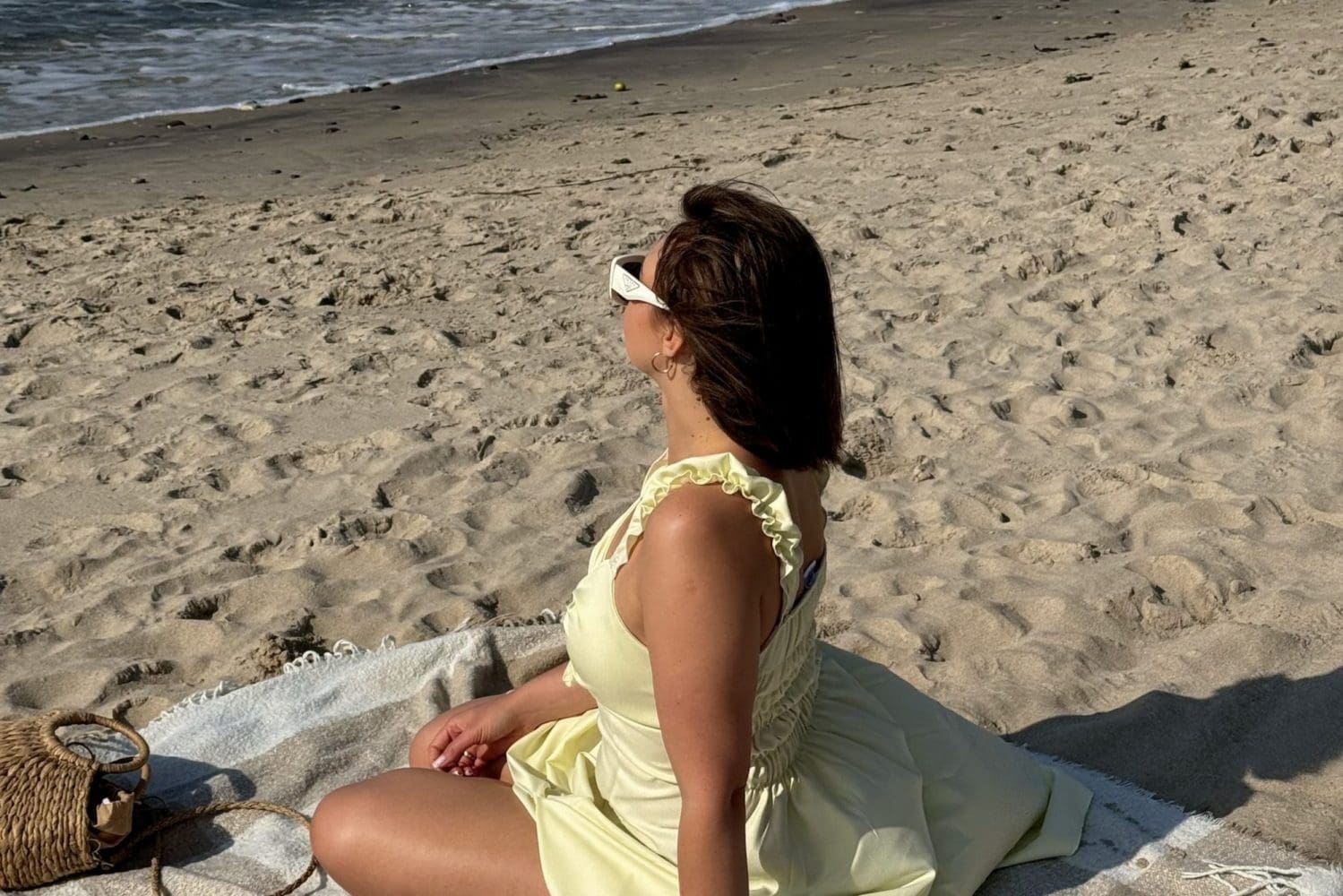 Kobieta w żółtej letniej sukience i okularach przeciwsłonecznych siedzi na kocu na słonecznej plaży nad Bałtykiem.