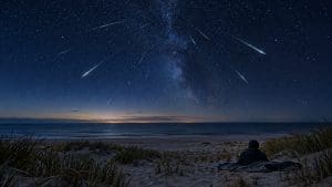 Smugi meteorów na gwiaździstym niebie nad ciemną plażą, mała postać na kocu patrzy w górę