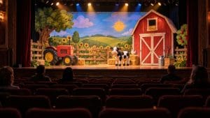 Kolorowa scenografia wiejskiego podwórka na małej scenie teatru, ciepłe reflektory, widownia widziana od tyłu