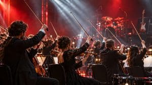 Skrzypkowie orkiestry unoszą smyczki w czerwono-białych światłach rockowego koncertu, w tle perkusja i gitara