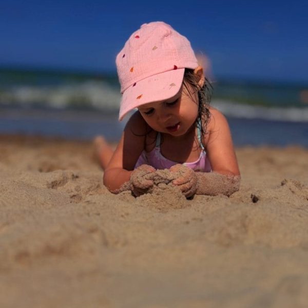 Dziewczynka w różowej czapce bawi się piaskiem na plaży, w tle morze