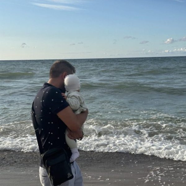 Tata z maluchem na rękach patrzy na fale nad brzegiem morza
