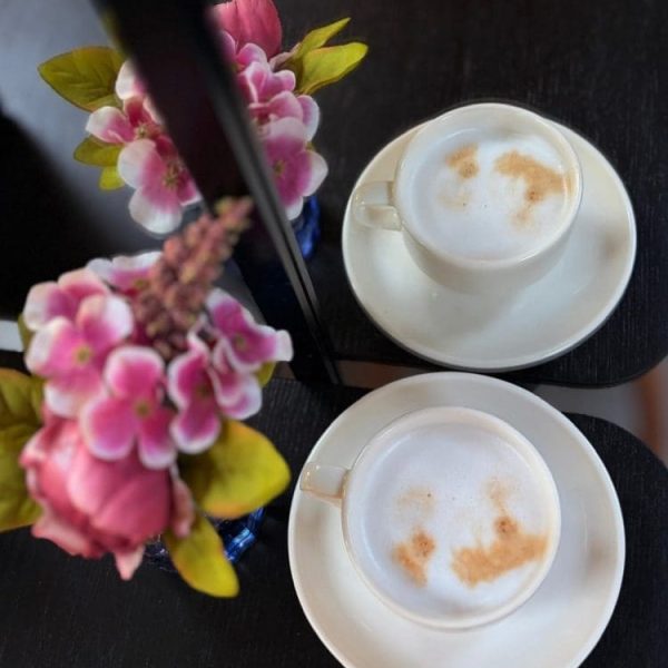 Dwie filiżanki cappuccino na stoliku, obok wazon z różowymi kwiatami