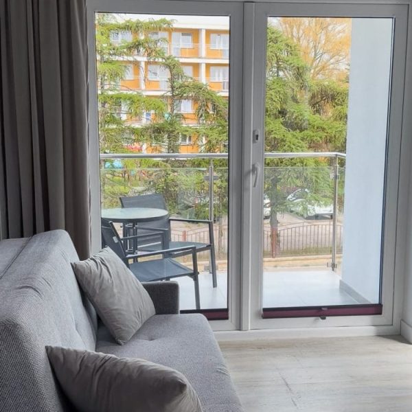Salon apartamentu z sofą i wyjściem na balkon, za szybą wiosenna zieleń